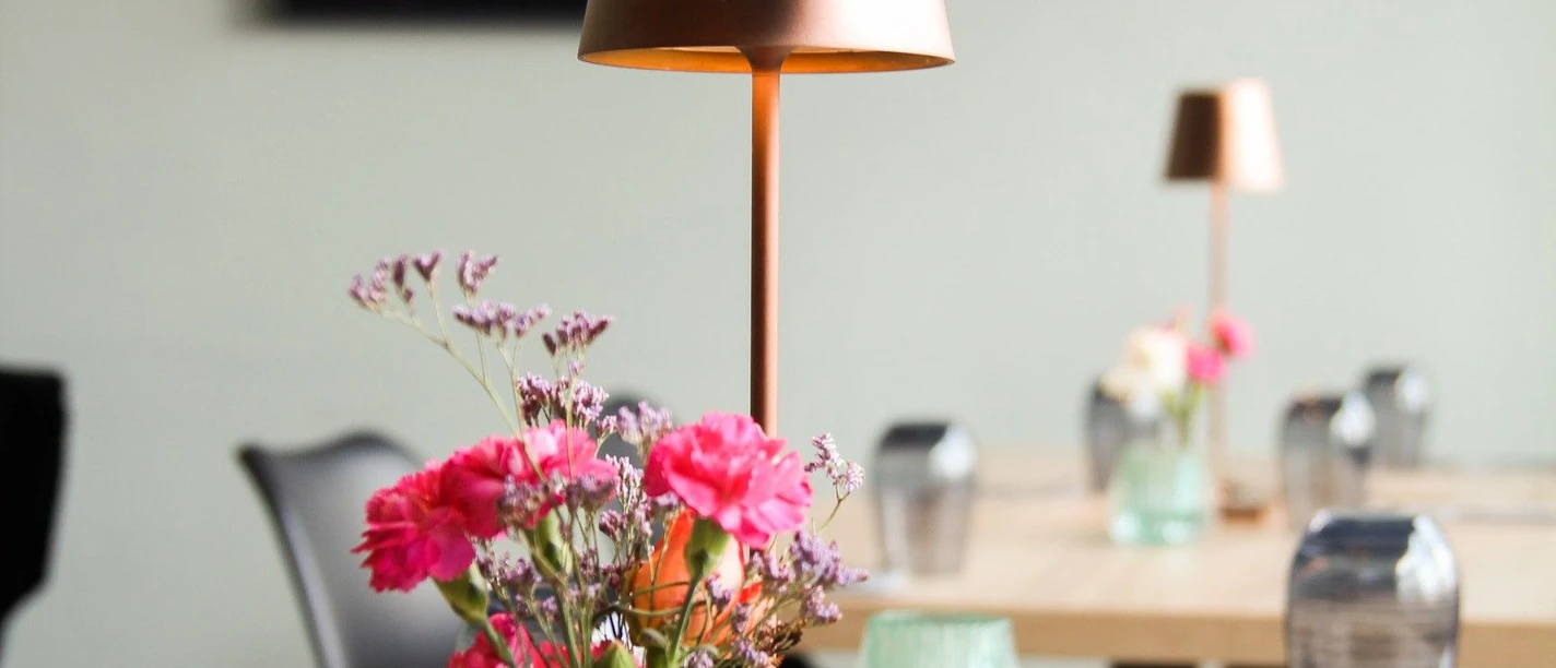 Tisch mit Gläsern, Teelicht, Vase mit Blumen, Besteck, Servietten und einer kleinen Stehlampe. Im Hintergrund hängt ein Bild an der Wand.