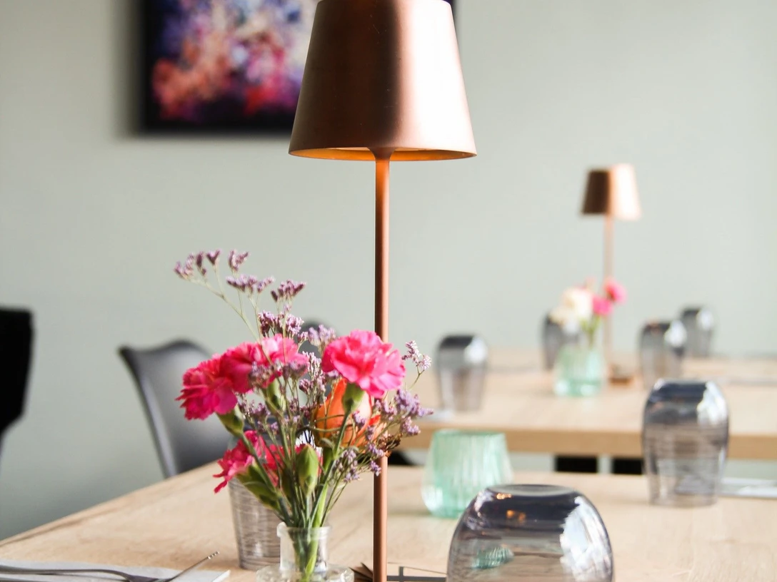 Trattoria Bar Centrale 46 (1).jpg Tisch mit Gläsern, Teelicht, Vase mit Blumen, Besteck, Servietten und einer kleinen Stehlampe. Im Hintergrund hängt ein Bild an der Wand.