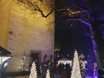 Weihnachtlich beleuchteter Kirchhof mit weißen Lichtkegeln, Menschen schlendern bei abendlicher Stimmung.