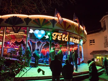 Weihnachtsmarkt - Feldmanns Raupe.jpg Ein belebter Jahrmarkt mit hell beleuchteten Karussells und zahlreichen Besuchern in der Abenddämmerung.