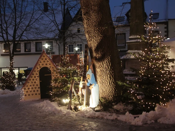 Weihnachtlich beleuchtete Parkszene mit Krippe, Tannenbäumen und schneebedecktem Boden bei Dämmerung.