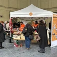 Eine Gruppe von Menschen steht vor einem Zelt mit Weihnachtsschmuck und einem „Weihnachtstausch beim Dekotausch“-Banner, beim Elisenbrunnen in Aachen.