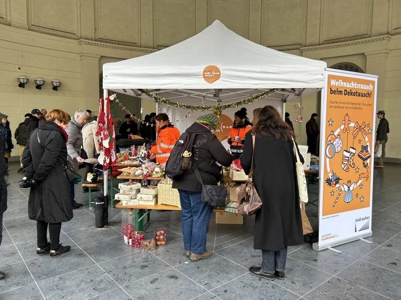Weihnachtsdekotausch Elisenbrunnen_(c)Stadt Aachen.jpg Eine Gruppe von Menschen steht vor einem Zelt mit Weihnachtsschmuck und einem „Weihnachtstausch beim Dekotausch“-Banner, beim Elisenbrunnen in Aachen.