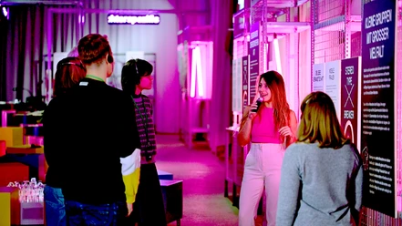 Eine Person führt eine Gruppe in einer Ausstellung mit rosa beleuchteten Tafeln und Exponaten.