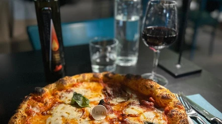 Pizza_Mozzarella_CCBYSA_Teutoburger Wald_Stadt_Schloß Holte-Stukenbrock.jpg Eine Holzofenpizza mit Mozzarella und frischem Basilikum, begleitet von Rotwein und Wasser auf einem Tisch.