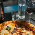 Pizza_Mozzarella_CCBYSA_Teutoburger Wald_Stadt_Schloß Holte-Stukenbrock.jpg Eine Holzofenpizza mit Mozzarella und frischem Basilikum, begleitet von Rotwein und Wasser auf einem Tisch.
