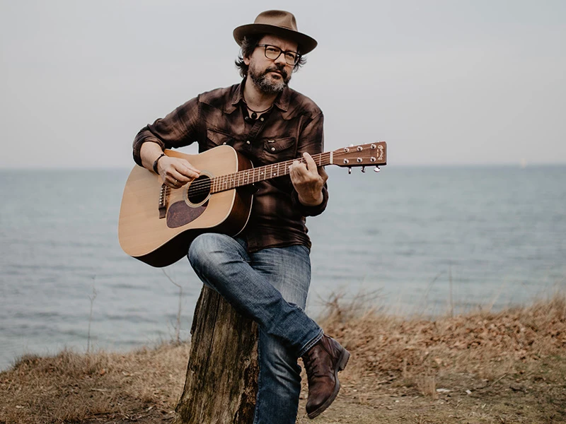 Trailhead_800_FotoRicoBurnecki.jpg Mann mit Hut spielt Akustikgitarre auf einem Baumstumpf am Seeufer, umgeben von grauem Himmel.
