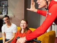 Ein Radfahrer in roter Kleidung spricht begeistert mit lächelnden Menschen auf einem gelben Sofa.