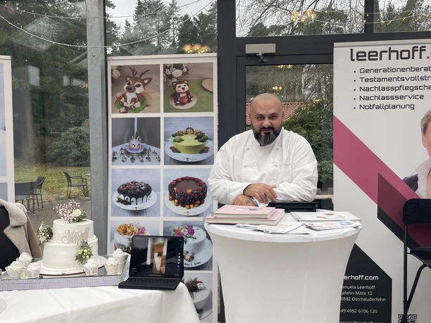 Konditor präsentiert Hochzeitstorten an Messestand mit Prospekten, Laptop und dekorierten Kuchen.