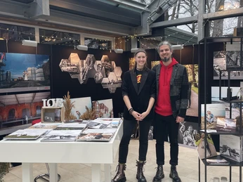 IMG_1965.jpg Zwei Personen stehen lächelnd in einer modernen Ausstellungshalle mit Fotografien und Broschüren.