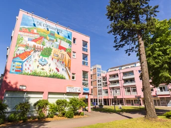 Großes Wandgemälde an einem rosa Wohngebäude, Bäume im Vordergrund, blauer Himmel.