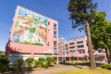 Espelkamp Großes Wandgemälde an einem rosa Wohngebäude, Bäume im Vordergrund, blauer Himmel.