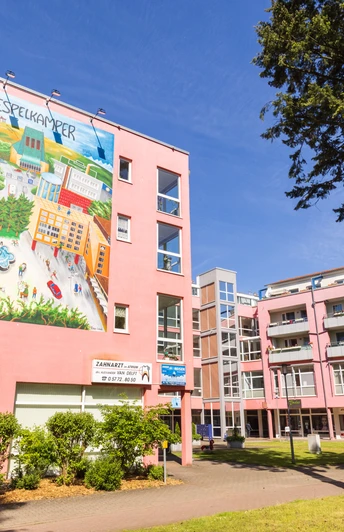 Espelkamp Großes Wandgemälde an einem rosa Wohngebäude, Bäume im Vordergrund, blauer Himmel.