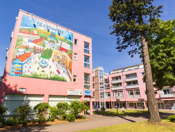 Espelkamp Großes Wandgemälde an einem rosa Wohngebäude, Bäume im Vordergrund, blauer Himmel.