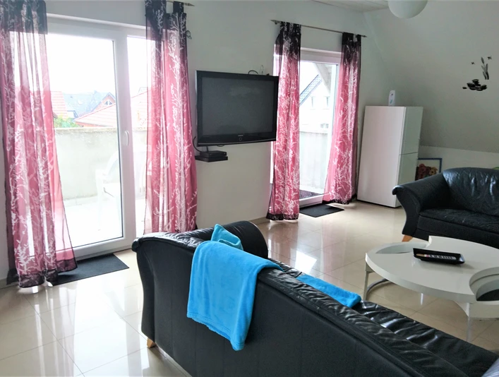 FeWo am Klinikum_Wohnzimmer-B Pipa.jpg Helles Wohnzimmer mit großen Fenstern, schwarzen Sofas, modern eingerichtet und Balkonzugang.