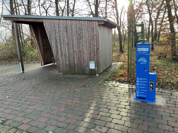 Radstation Mitte Niedersachsen Eine Fahrradstation mit blauer Reparatursäule steht auf einem gepflasterten Weg vor einem hölzernen Unterstand.