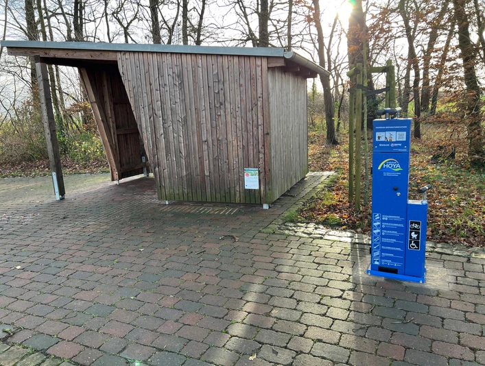 Radstation Mitte Niedersachsen Eine Fahrradstation mit blauer Reparatursäule steht auf einem gepflasterten Weg vor einem hölzernen Unterstand.