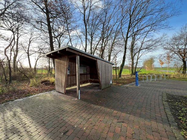 Radstation Mitte Niedersachsen Überdachter Rastplatz an einem Radweg mit Holzverkleidung, Fahrradständer und umgeben von Bäumen.