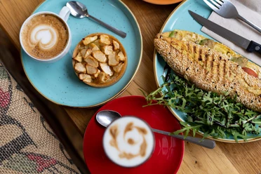 Auf einem Holztisch liegen ein Cappuccino mit Herzmotiv und eine Schale mit Apfelkuchen, neben einem belegten Baguette mit Salatgarnitur.