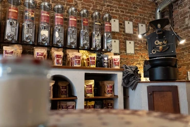 Innenbereich eines Cafés mit Regalen voller Kaffeebohnenbehälter und einem traditionellen Kaffeeröster an einer Backsteinwand.