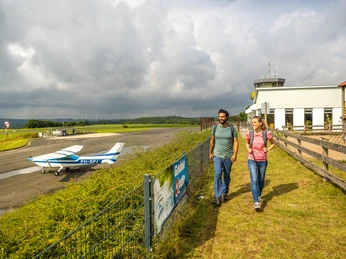 Höxter-Flugplatz Höxter-Holzminden-Teutoburger-Wald-Stadt Höxter-D-Ketz-016.jpg Wanderndes Pärchen vor kleinem Sportflugzeug in Parkposition mit Landebahn und Flughafengebäude im HintergrundWalking couple in front of a small sports airplane in parking position with runway and airport building in the background
