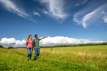 Über Wiesen wanderndes Pärchen mit hoch getürmten Wolken im HintergrundCouple walking across meadows with towering clouds in the background