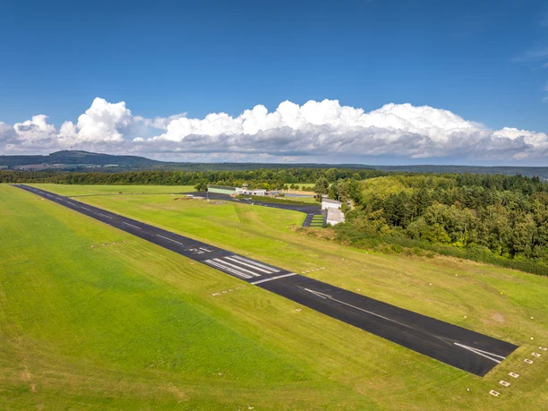 Aerial view of the runway surrounded by mown meadow and landscape with airport building and hangar on the right of the picture