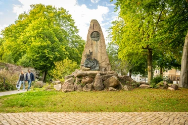Höxter-Hoffmann von Fallersleben Denkmal-Teutoburger-Wald-Stadt Höxter-D-Ketz-165.jpg Denkmal aus Naturstein mit integriertem Bronzekopf von Hoffmann von Fallersleben mit Bäumen und zwei Spaziergängern