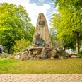 Höxter-Hoffmann von Fallersleben Denkmal-Teutoburger-Wald-Stadt Höxter-D-Ketz-165.jpg Denkmal aus Naturstein mit integriertem Bronzekopf von Hoffmann von Fallersleben mit Bäumen und zwei SpaziergängernNatural stone monument with integrated bronze head by Hoffmann von Fallersleben with trees and two walkers
