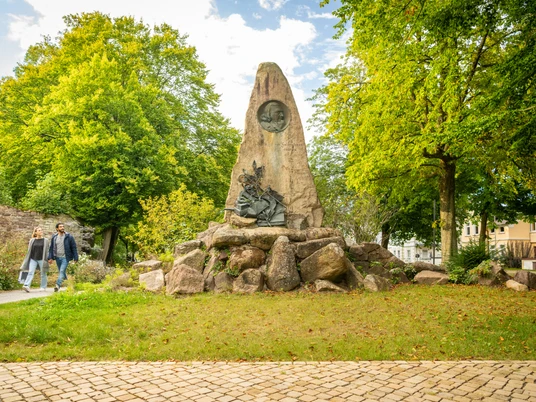 Höxter-Hoffmann von Fallersleben Denkmal-Teutoburger-Wald-Stadt Höxter-D-Ketz-165.jpg Denkmal aus Naturstein mit integriertem Bronzekopf von Hoffmann von Fallersleben mit Bäumen und zwei SpaziergängernNatural stone monument with integrated bronze head by Hoffmann von Fallersleben with trees and two walkers