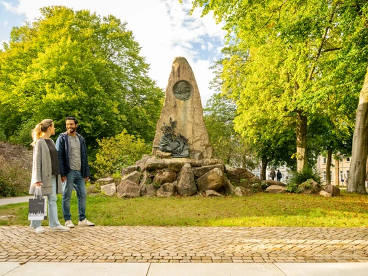Höxter-Hoffmann von Fallersleben Denkmal-Teutoburger-Wald-Stadt Höxter-D-Ketz-166.jpg Denkmal aus Naturstein mit integriertem Bronzekopf von Hoffmann von Fallersleben mit Bäumen und zwei SpaziergängernNatural stone monument with integrated bronze head by Hoffmann von Fallersleben with trees and two walkers