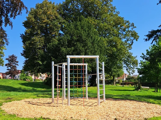1_ParkMitPotential_quer.jpg Klettergerüst aus Metall auf Holzboden, umgeben von grünem Rasen und Schatten spendenden Bäumen.