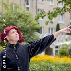 Eine Person in historischer Kleidung steht vor einem Stadtgebäude und zeigt auf etwas im Off.