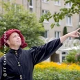 Eine Person in historischer Kleidung steht vor einem Stadtgebäude und zeigt auf etwas im Off.