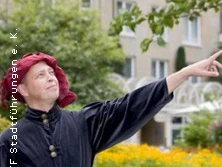 Eine Person in historischer Kleidung steht vor einem Stadtgebäude und zeigt auf etwas im Off.
