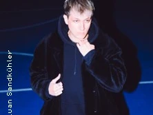 Eine Person in dunkler Kleidung auf blauer Fläche mit nachdenklichem Blick und einer Hand am Kinn.