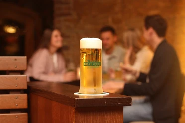 Barres Brauwelt - Frisch gezapft Ein frisch gezapftes Glas Bier steht im Vordergrund, mehrere Personen in geselliger Runde im Hintergrund.