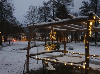 Verschneiter Stadtpark bei Dämmerung, hölzerner Pavillon mit leuchtenden Lichterketten geschmückt.