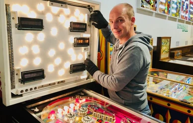 Technikführung Ein Mann öffnet das leuchtende Kopfteil eines Flipperautomaten in einem Technikmuseum.