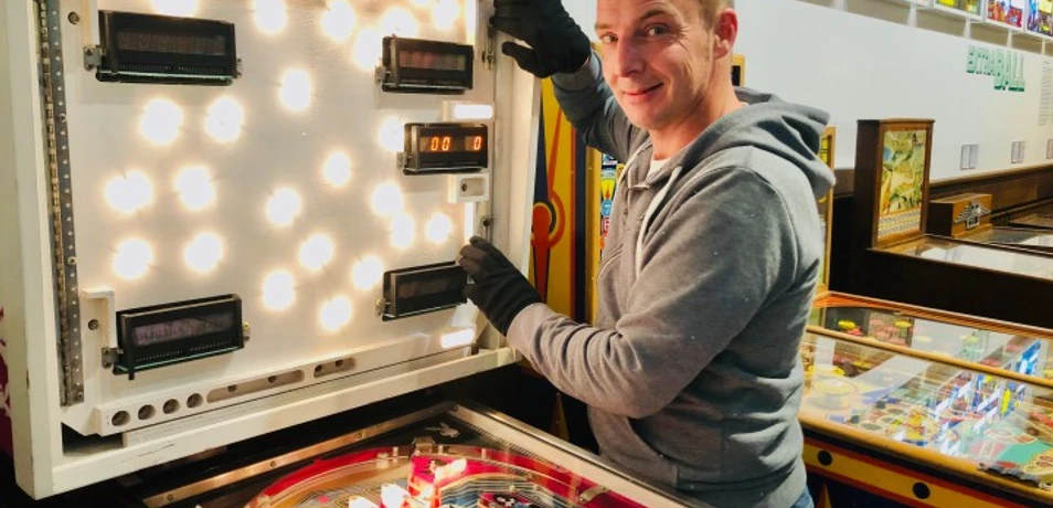 Technikführung Ein Mann öffnet das leuchtende Kopfteil eines Flipperautomaten in einem Technikmuseum.