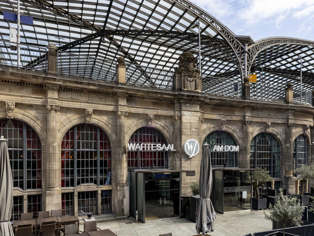 Wartesaal am Dom Historisches Gebäude mit prachtvollem Glasdach, davor Terrassenbereich; Aufschrift "Wartesaal".