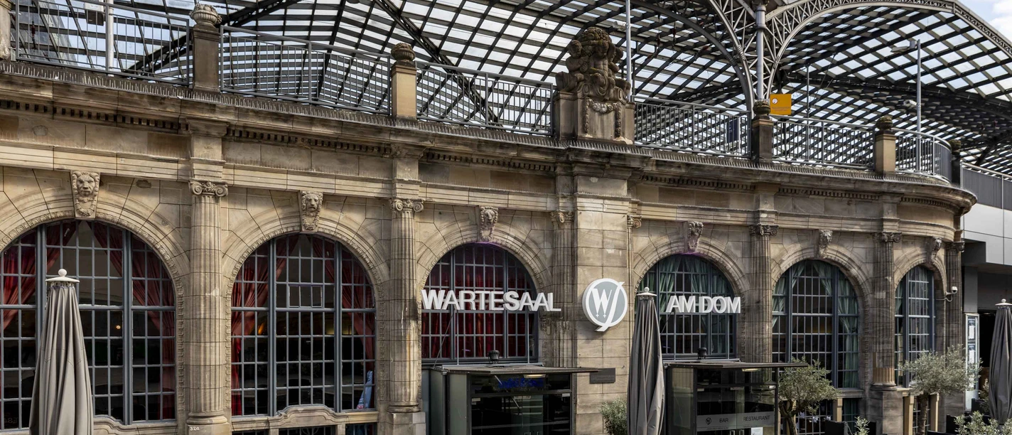 Wartesaal am Dom Historisches Gebäude mit prachtvollem Glasdach, davor Terrassenbereich; Aufschrift "Wartesaal".