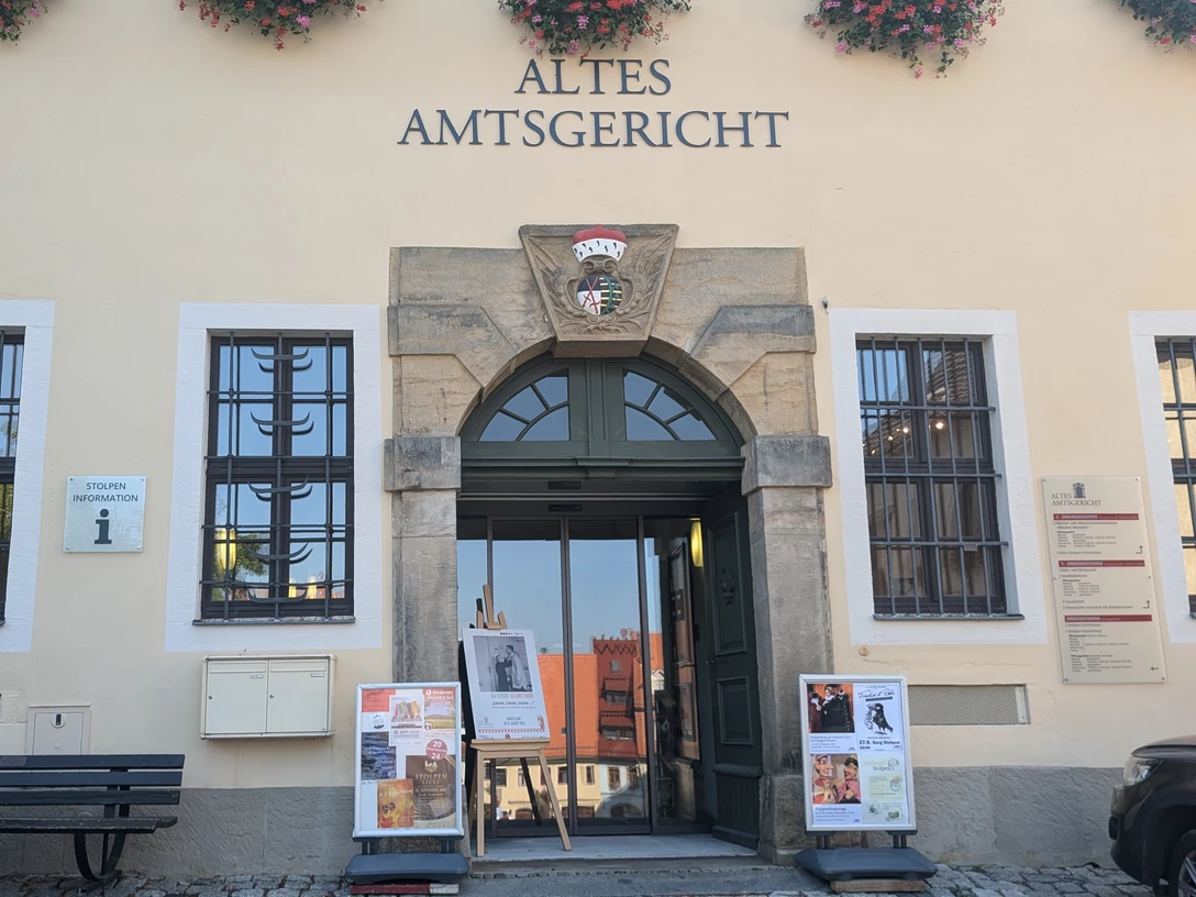 tourist-information-stolpen-(c)-heike-grunow-cc0.jpg Eingang des Alten Amtsgerichts mit Blumen über dem Schild, Plakate vor der Tür und einer Bank daneben; ruhige Atmosphäre.