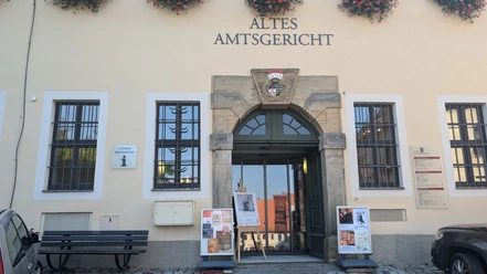 Eingang des Alten Amtsgerichts mit Blumen über dem Schild, Plakate vor der Tür und einer Bank daneben; ruhige Atmosphäre.