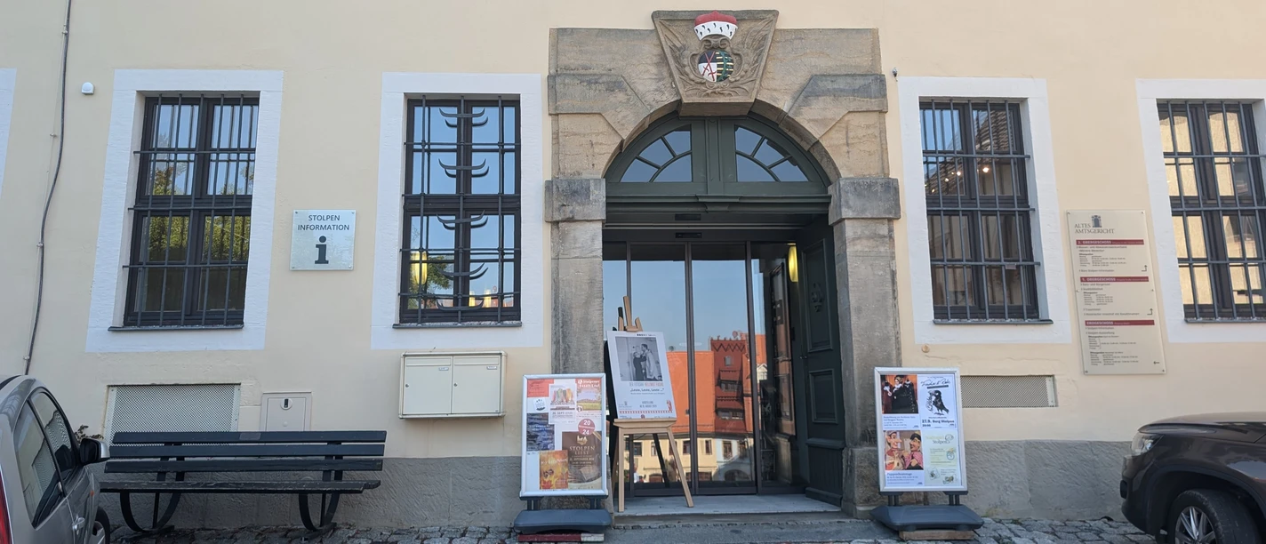 Eingang des Alten Amtsgerichts mit Blumen über dem Schild, Plakate vor der Tür und einer Bank daneben; ruhige Atmosphäre.