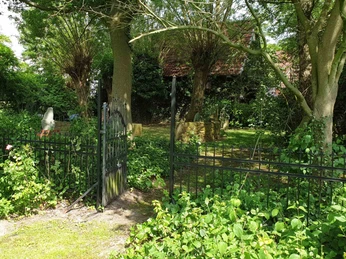 NaturGarten mit KunstObjekten -Gartentor zum alten Garten Metalltor im offenen Zustand eingerahmt von grünen Sträuchern, dahinter alter Garten mit Bäumen und Skulpturen.