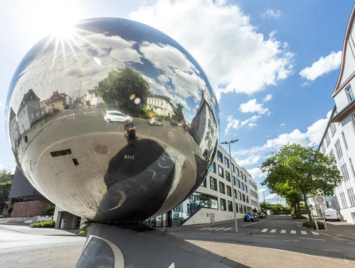 Kunstwerk aus Edelstahl reflektiert urbane Umgebung in Herford unter blauem Himmel mit weißen Wolken.