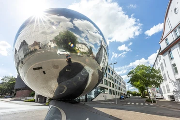 herford-marta-teutoburger-wald-tourismus-d-ketz-082-cc-by-sa-jpg.jpg Kunstwerk aus Edelstahl reflektiert urbane Umgebung in Herford unter blauem Himmel mit weißen Wolken.