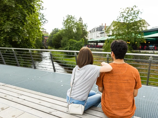 herford-promenade-teutoburger-wald-tourismus-h-tornow-003-jpg.jpg Ein Paar sitzt auf einer modernen Brücke am Fluss, umgeben von Bäumen und einem Café.