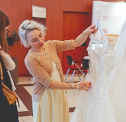 brautkleid.jpg Frau betrachtet ein detailreich besticktes weißes Brautkleid auf einem Kleiderbügel in Innenraum.